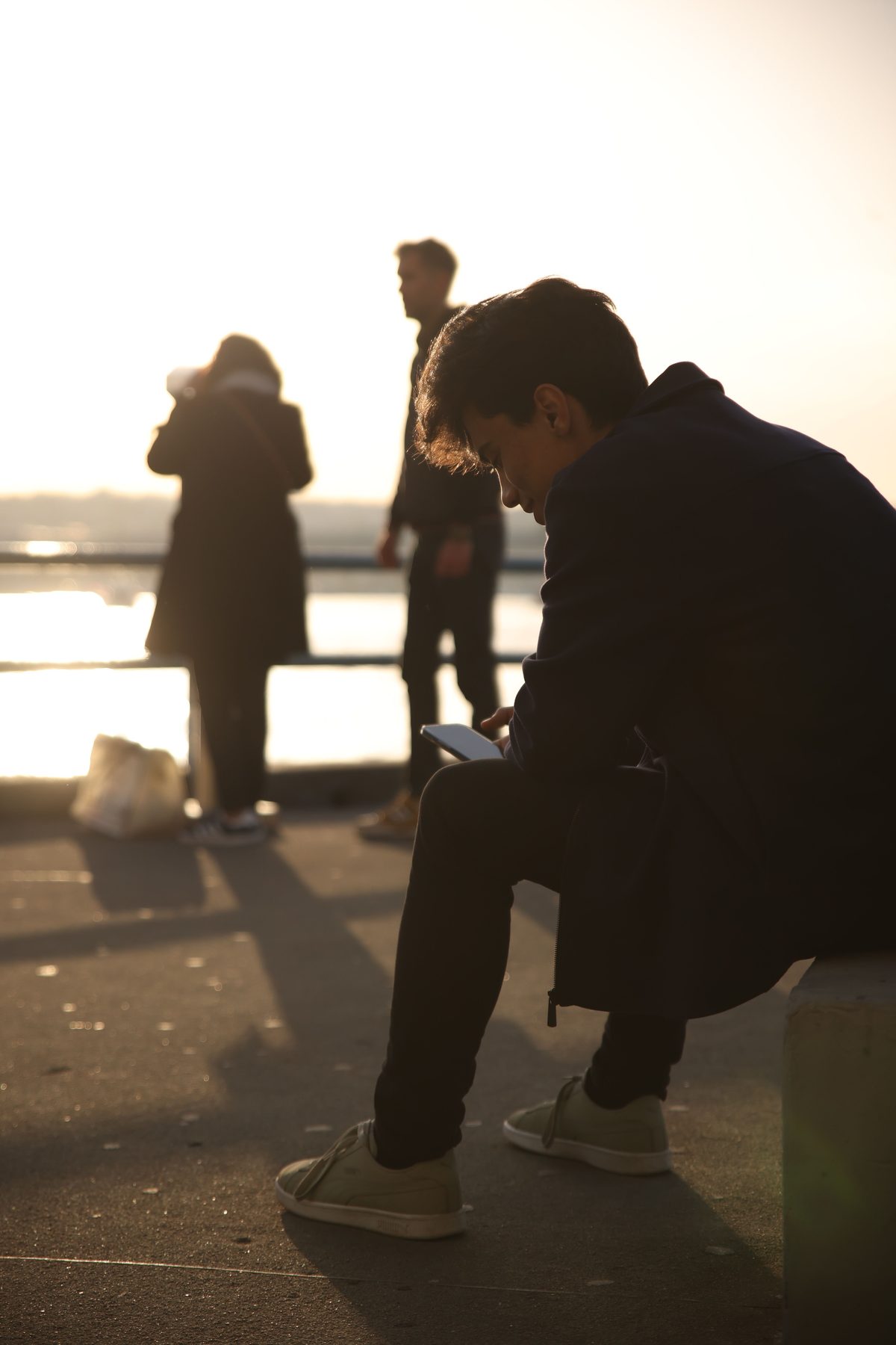 A young man sitting outdoors at sunset, reading his phone in warm golden light